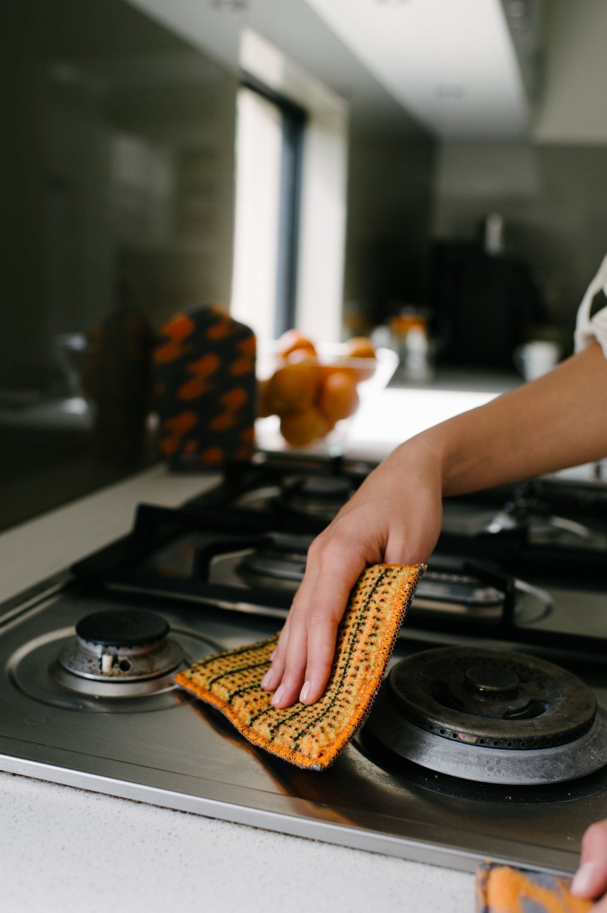 6 Aesthetic Cleaning Videos That Make Organizing Look Like Art 4 A hand wiping a stainless steel gas stovetop with a yellow textured sponge in a kitchen.