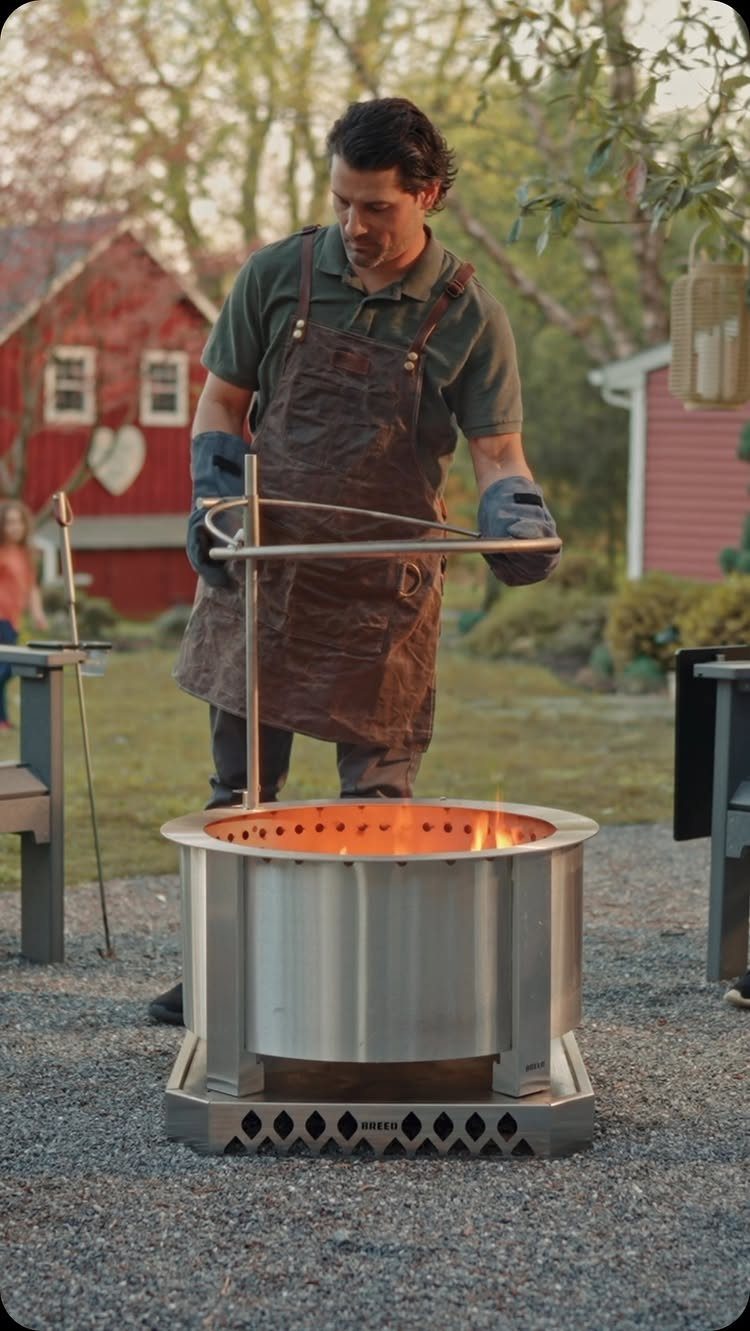 A man in an apron tending to a burning stainless steel fire pit outdoors.