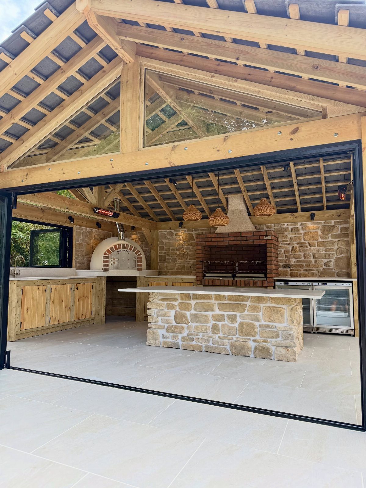 An outdoor kitchen with a stone island, brick grill, and pizza oven under a wooden pavilion.