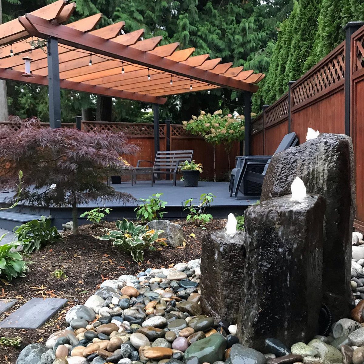 A dark stone water fountain bubbling amidst smooth river rocks and green garden plants.