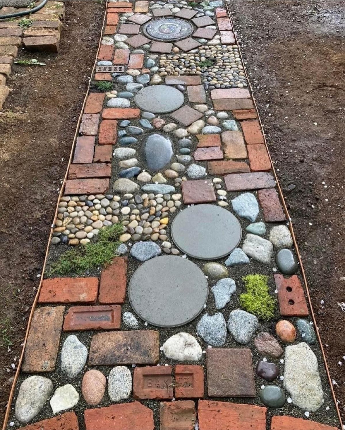 A decorative garden pathway composed of red bricks, smooth pebbles, and large circular grey stepping stones.