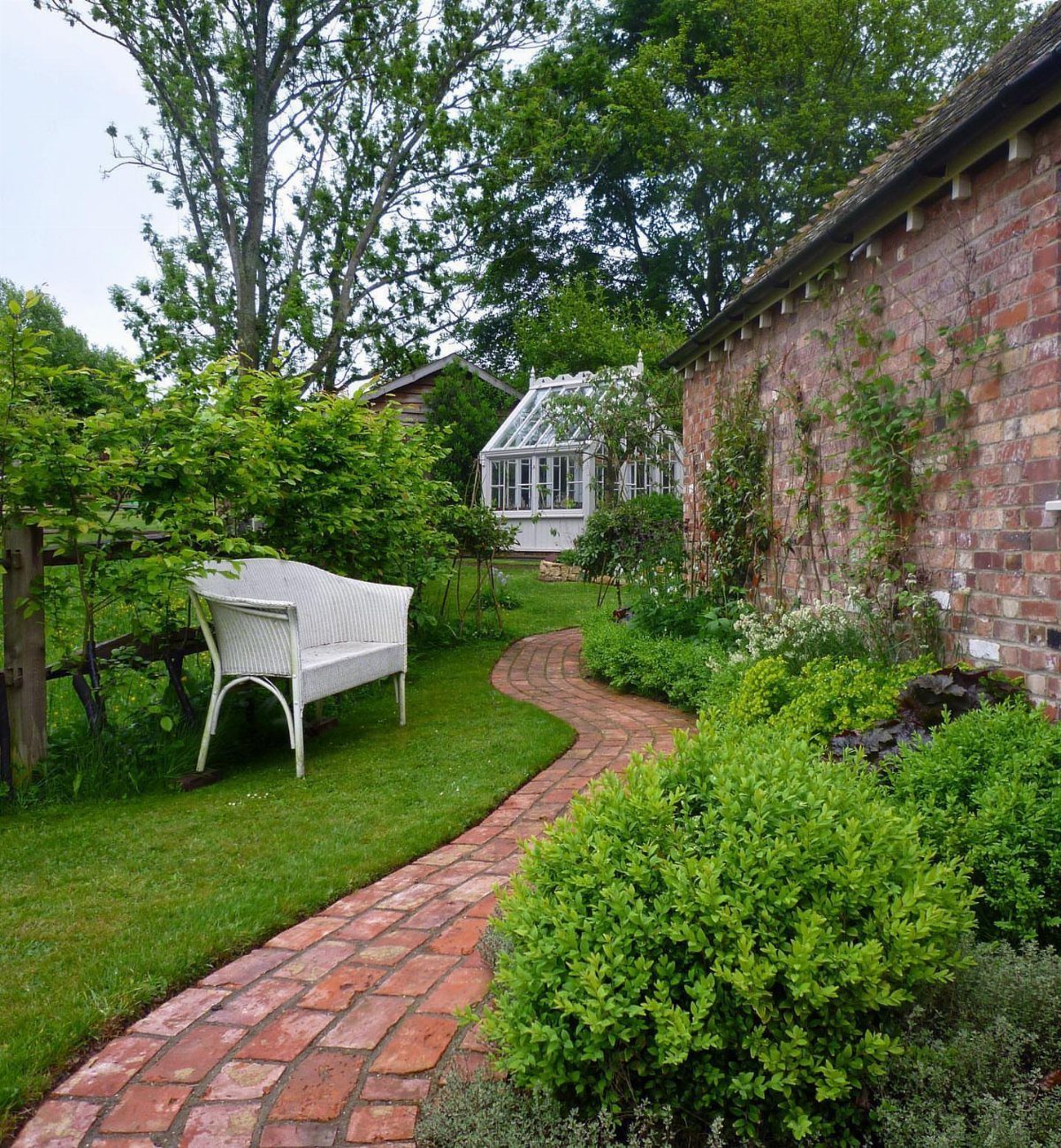 A curved red brick garden pathway winding through a lush green lawn and garden plants.