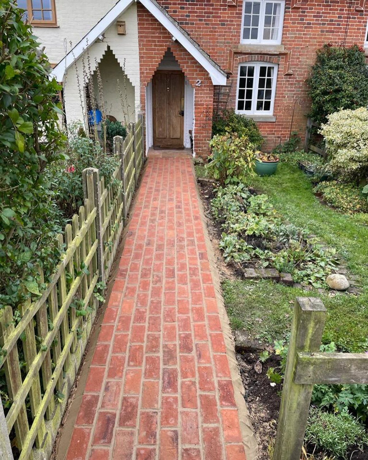 A straight red brick garden pathway leading toward a house entrance with a wooden door.