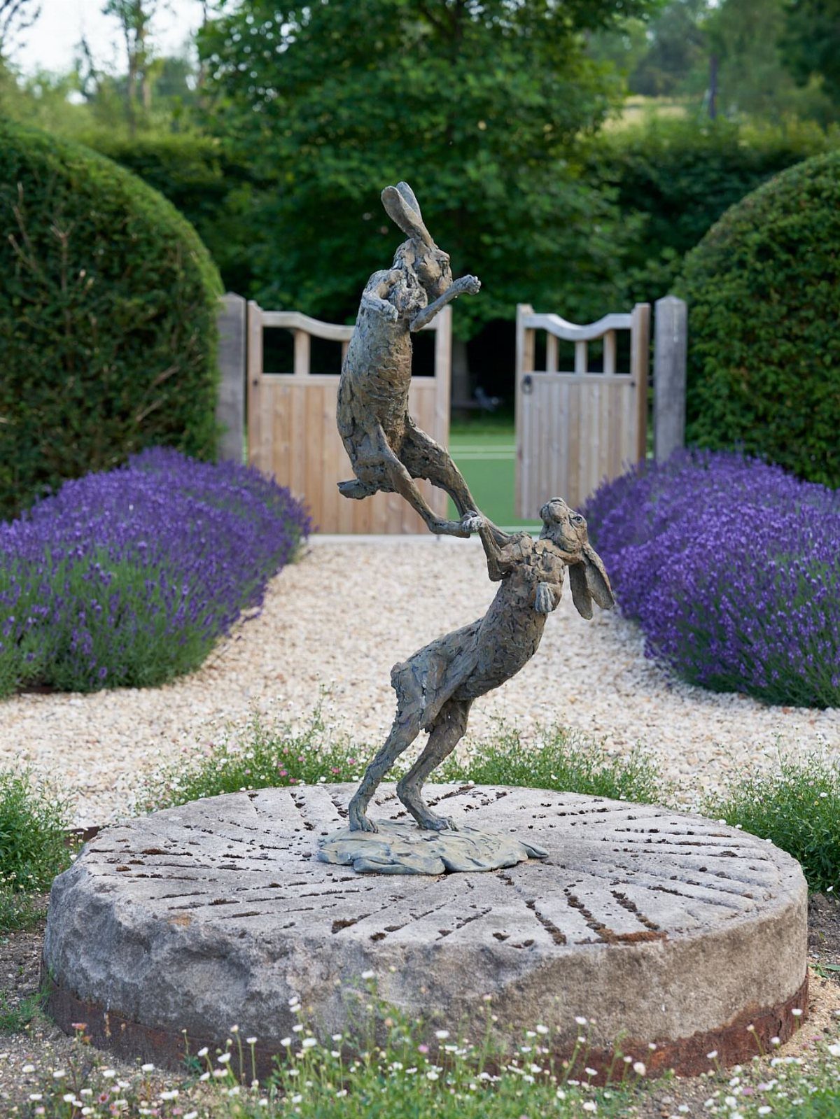 Bronze sculpture of two dancing hares on a stone plinth in a lavender garden.
