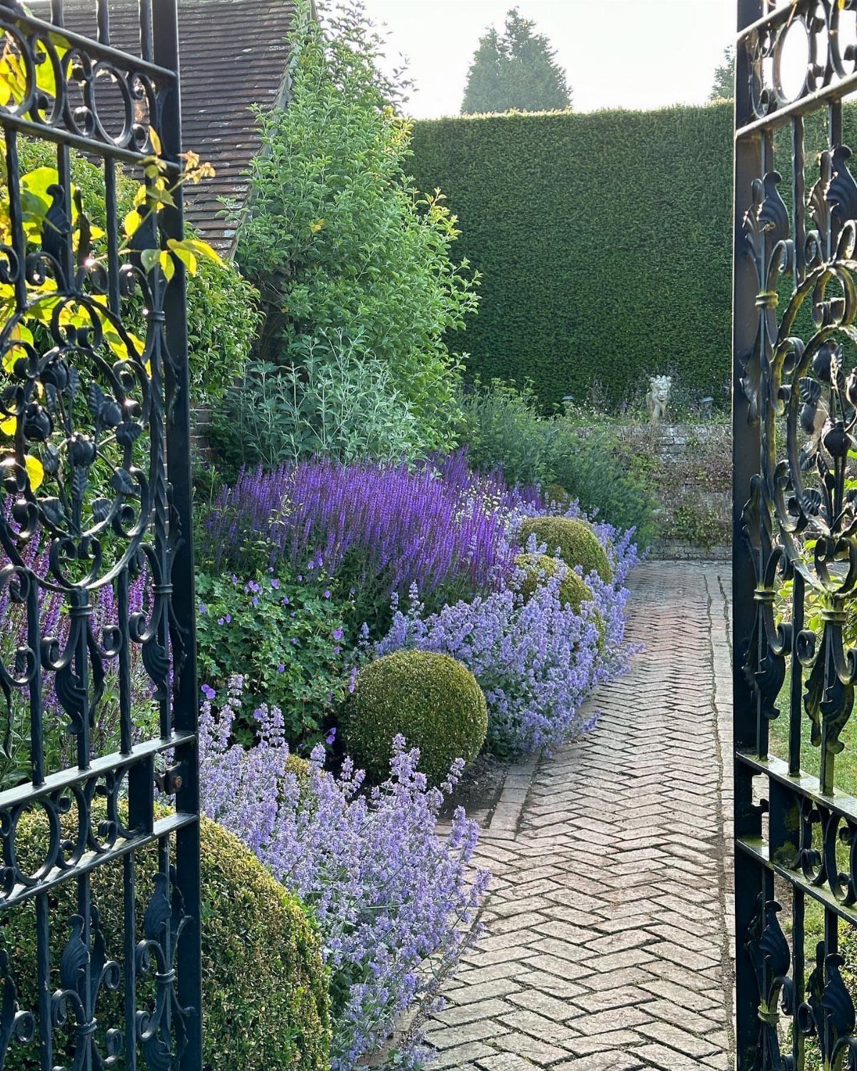 Purple flowers and green shrubs line a brick path viewed through an open iron gate.