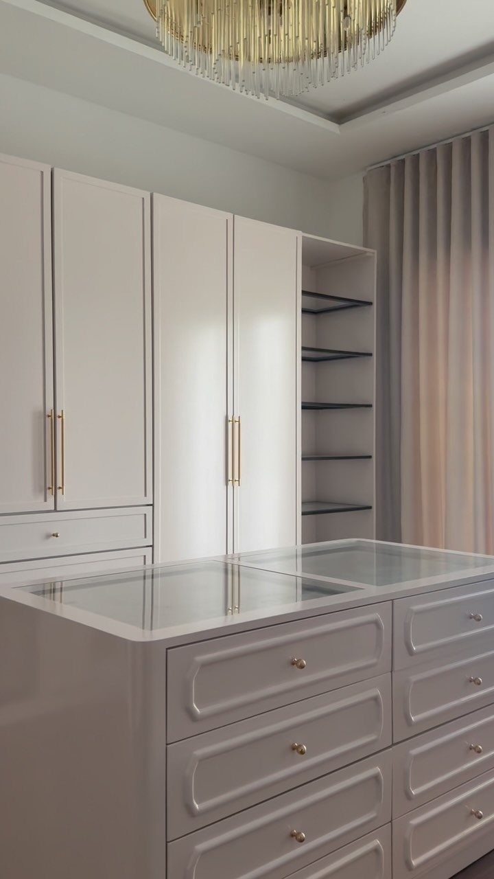 A light gray closet island with glass top and drawers sits in a modern walk-in closet.