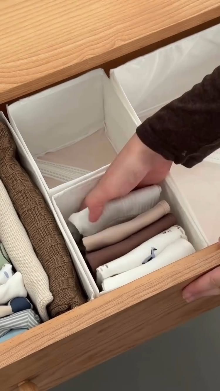 A hand organizing folded neutral-colored clothes inside white fabric drawer dividers within a wooden drawer.