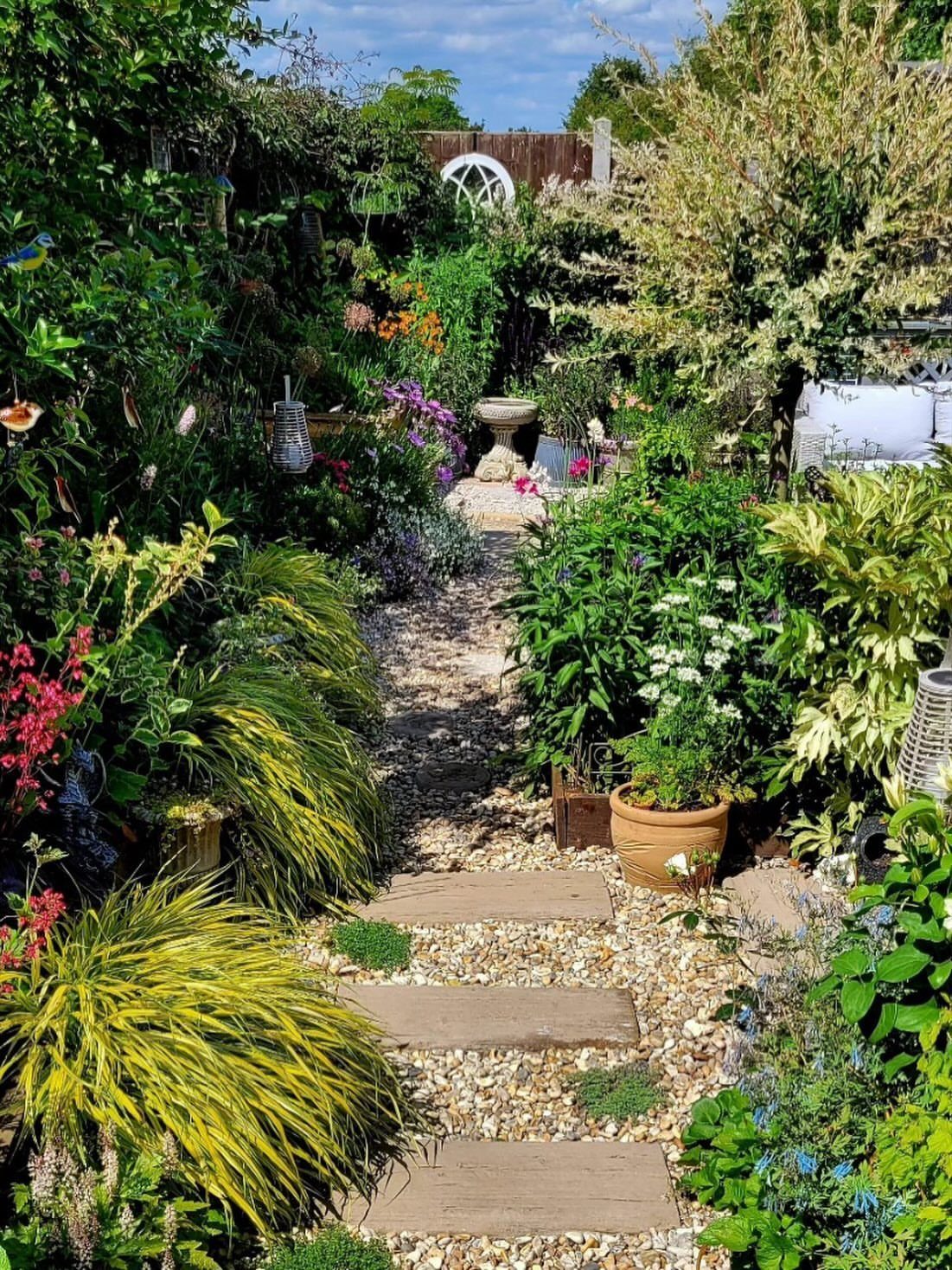 A gravel garden path with rectangular stone slabs winding through lush green plants and flowers.