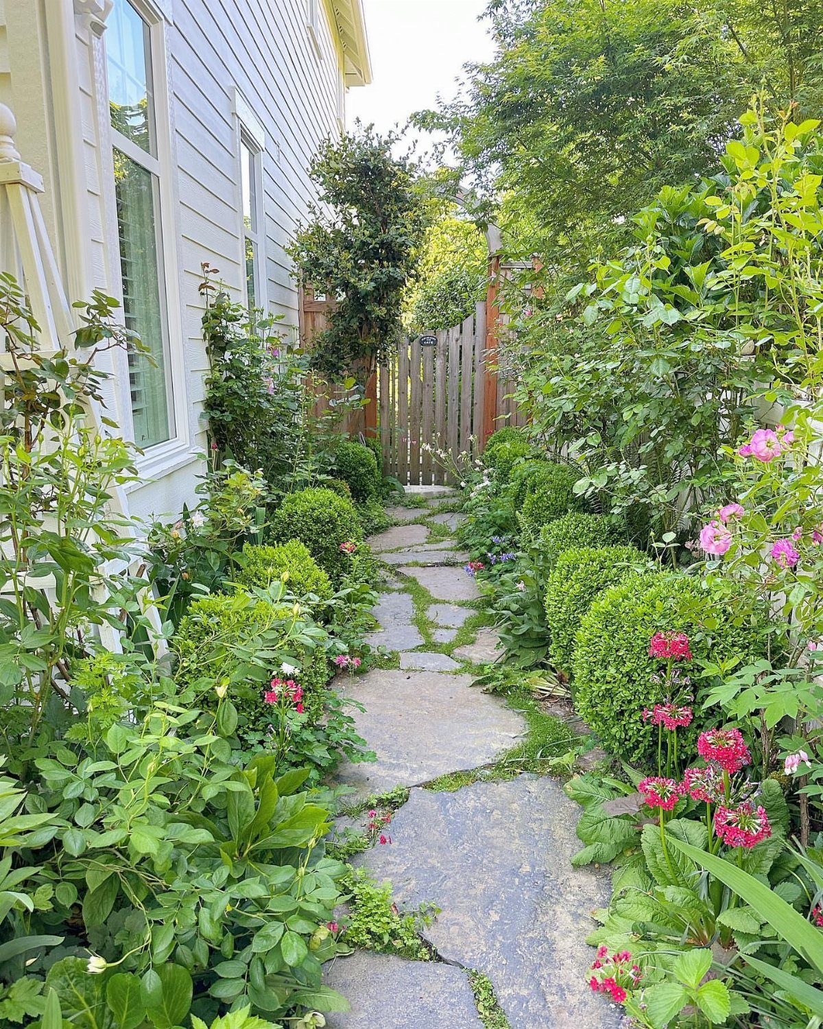 Irregular grey stone slabs form a garden pathway surrounded by lush green plants and pink flowers.