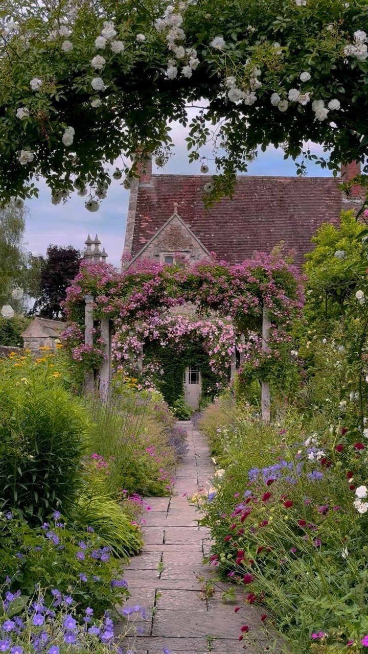 A stone garden path winding through colorful wildflowers toward a house with rose-covered arches.