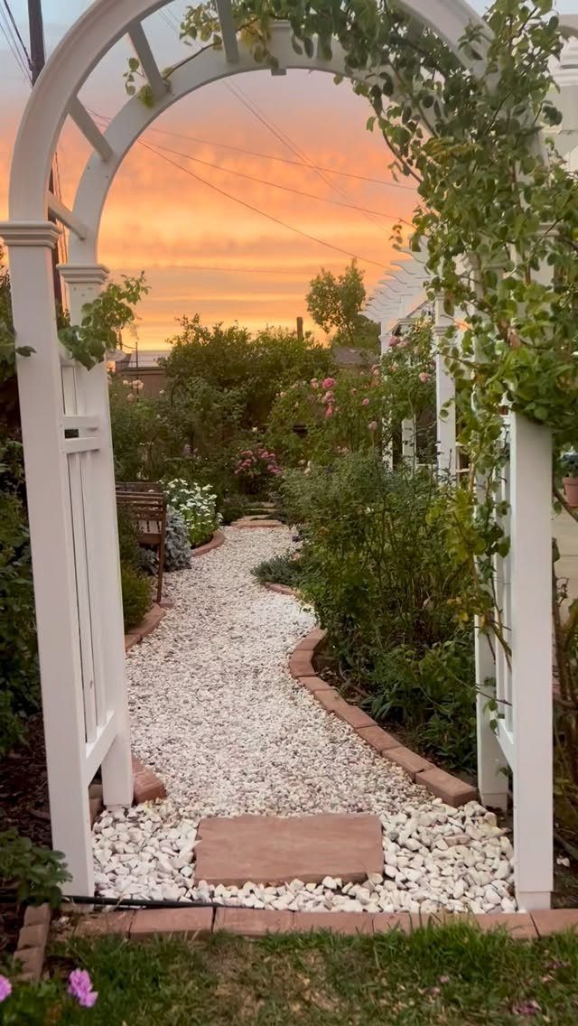 A white garden arch frames a winding white gravel path under a sunset sky.