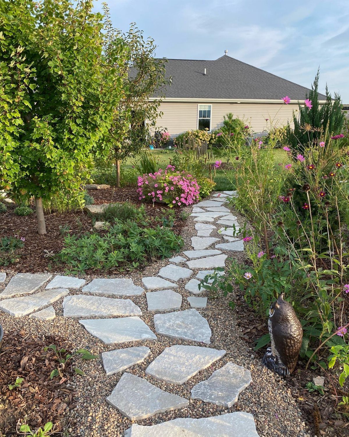 Irregular flagstone stepping stones create a curved garden path surrounded by green plants and pink flowers.
