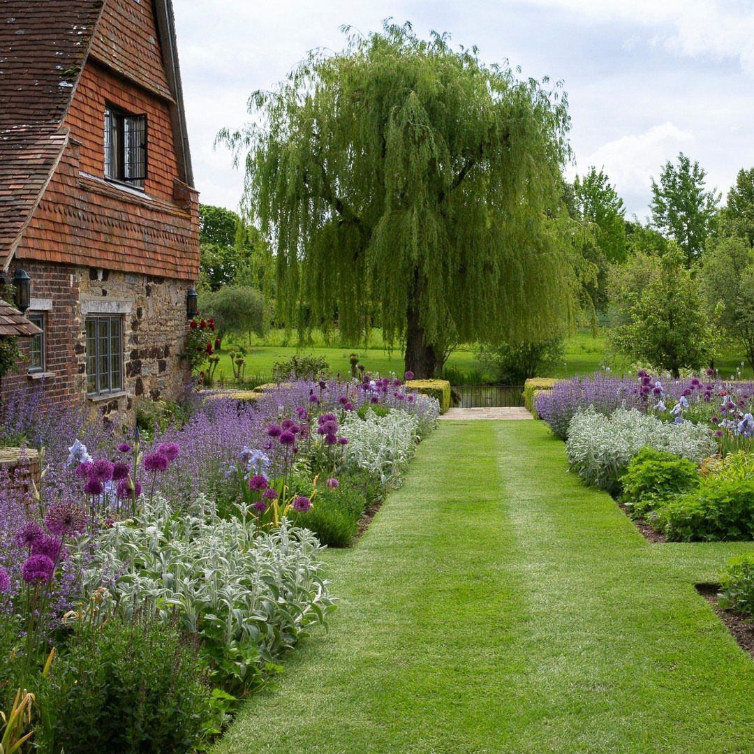 A manicured green grass path bordered by purple and silver flowers leading toward a willow tree.