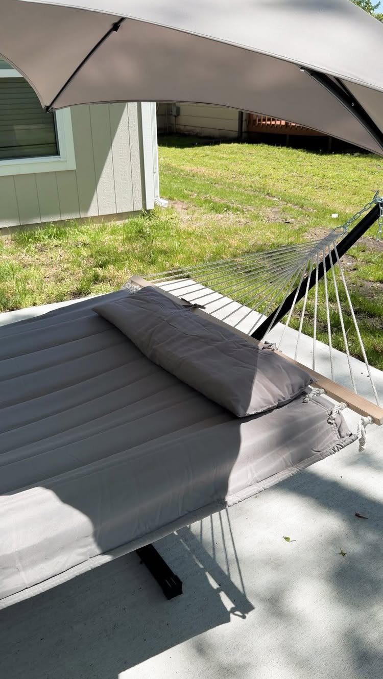 A grey fabric hammock with a pillow resting on a concrete patio under a large umbrella.