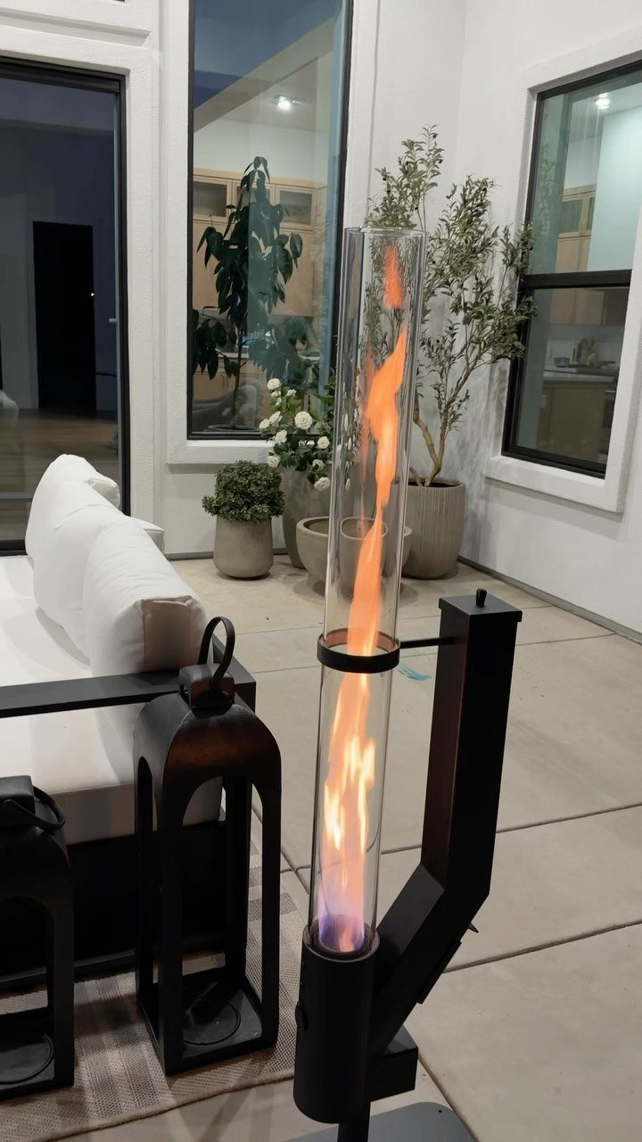 A tall glass flame torch with orange fire glowing inside on a modern patio.