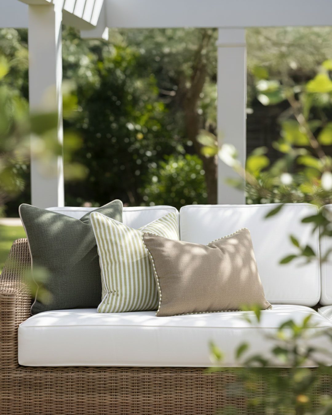 A wicker outdoor sofa with white cushions and decorative throw pillows in a sunny garden.