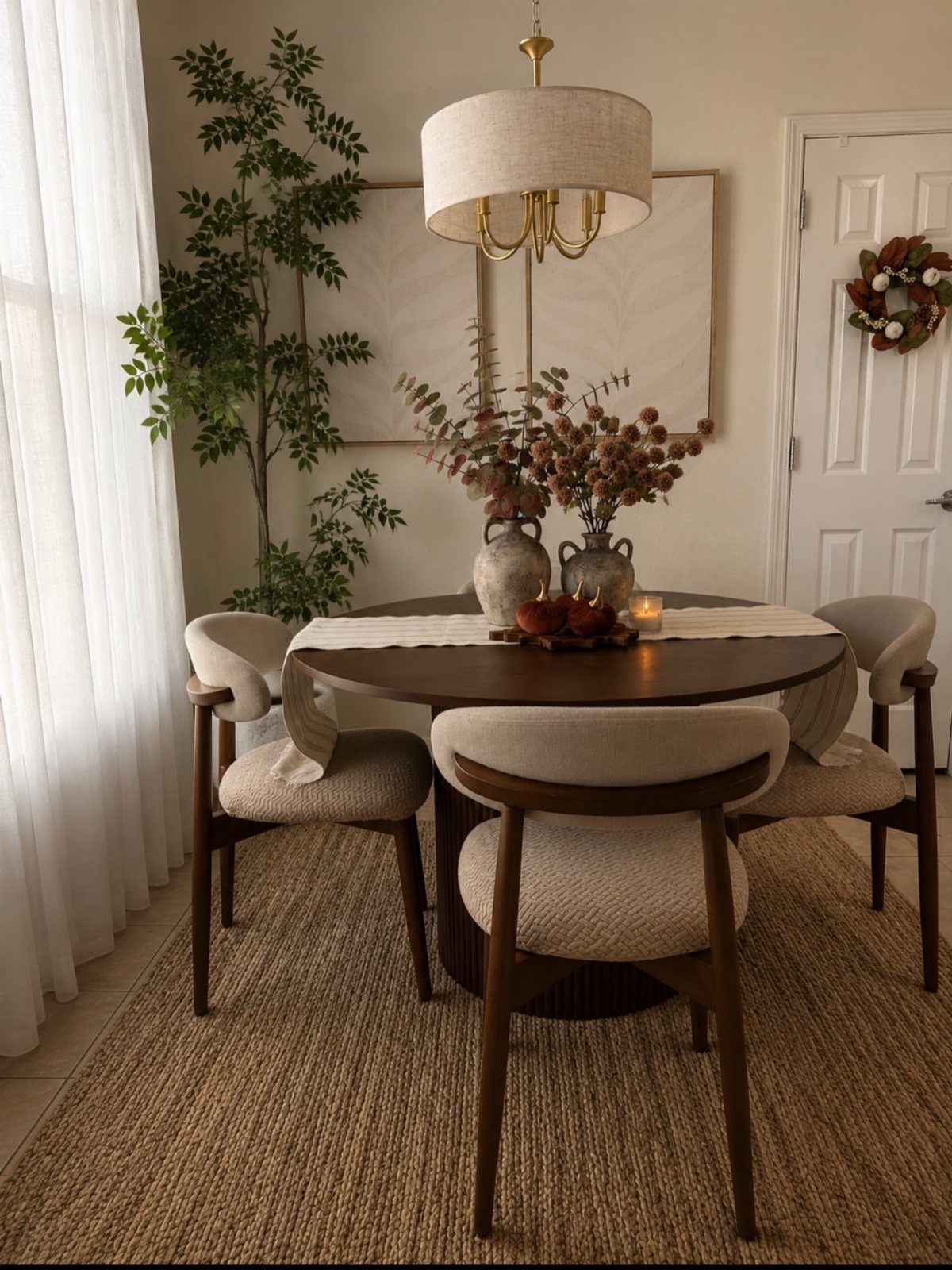 A round wooden dining table decorated with a white runner and floral vases in a bright room.
