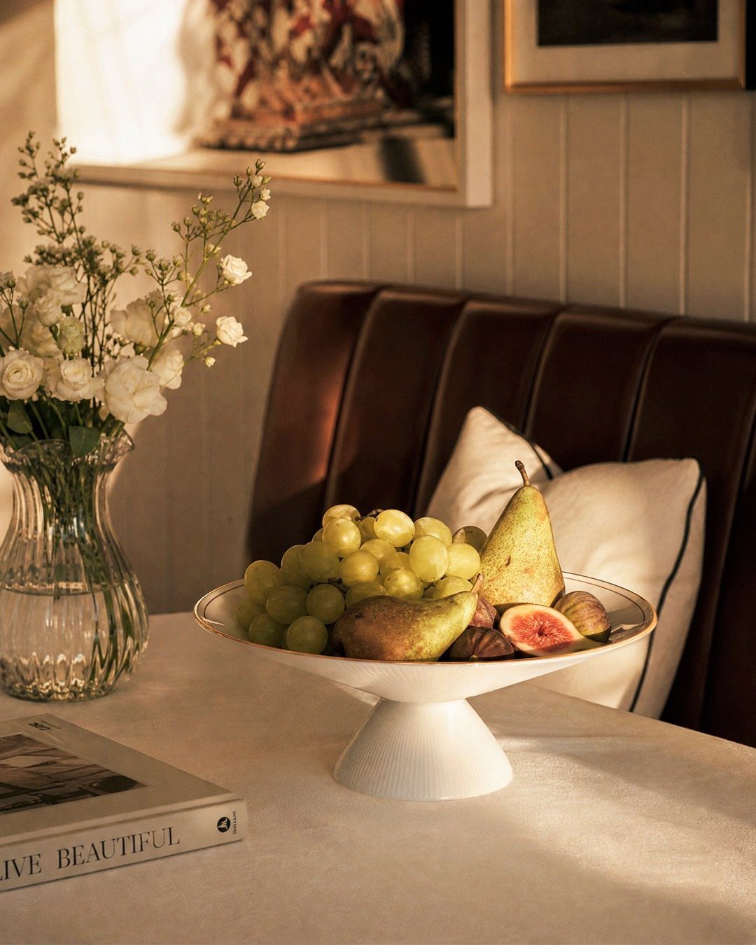 A white footed bowl filled with grapes, a pear, and figs on a dining table.