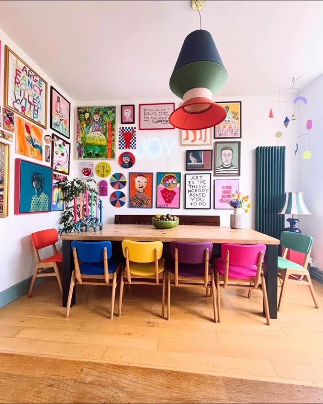 Colorful dining chairs arranged around a wooden table in a room with a large gallery wall.