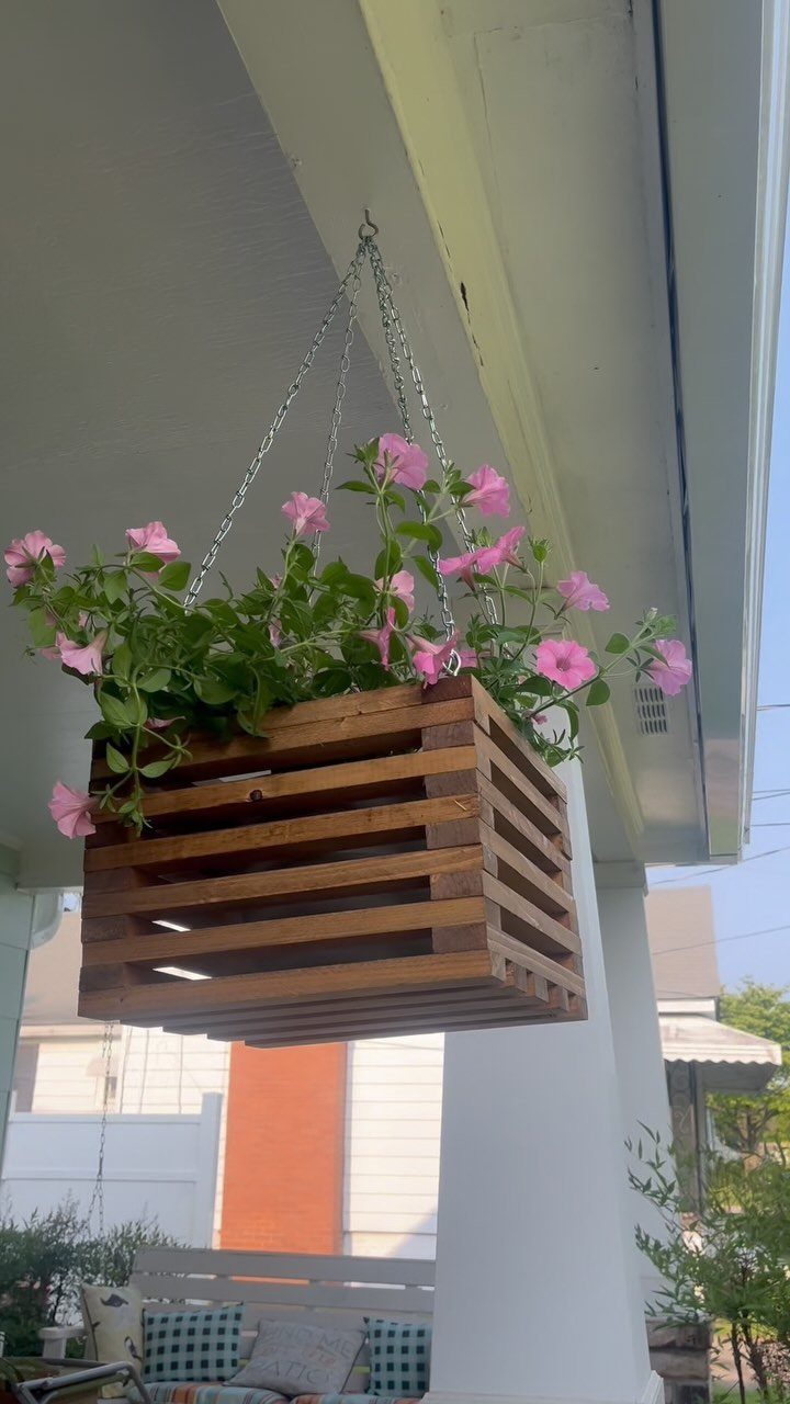 A wooden crate hanging basket filled with pink petunia flowers suspended by chains from a porch ceiling.
