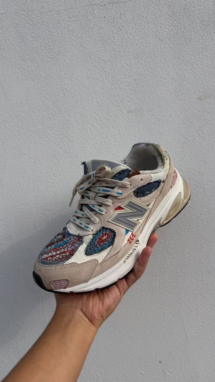 A hand holding a New Balance sneaker decorated with colorful fabric patterns and textures.