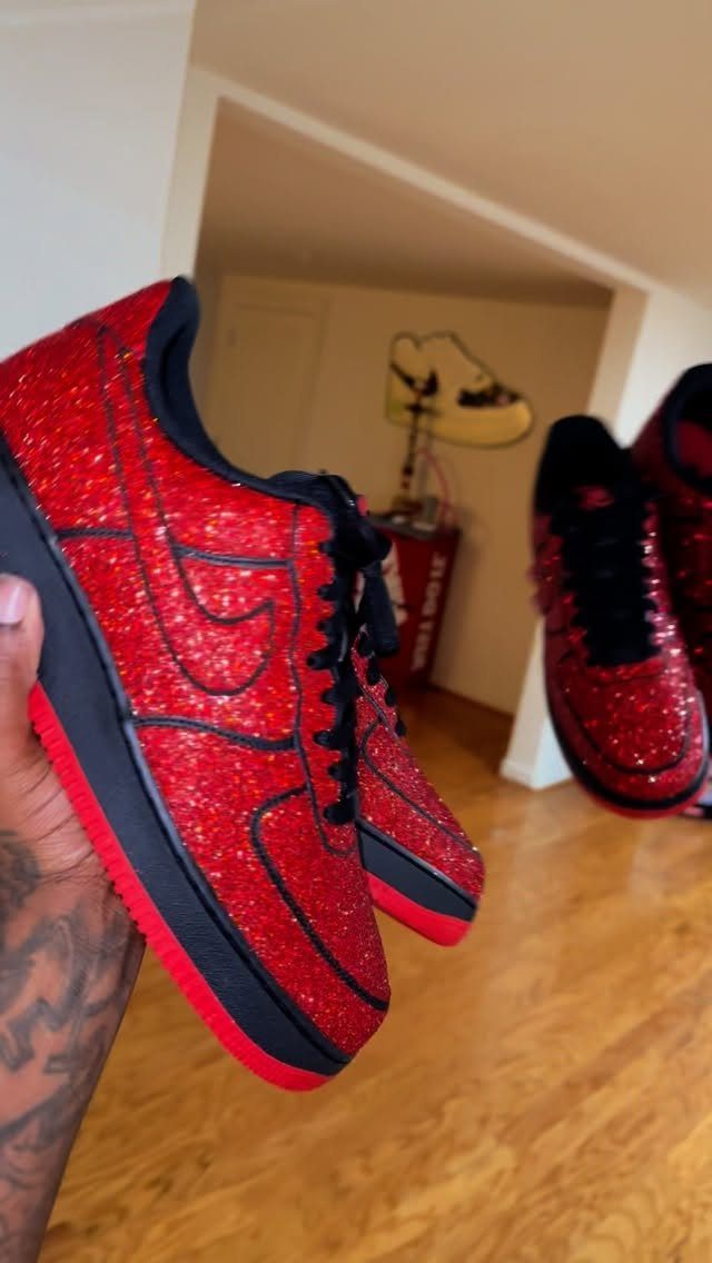 Red glitter-covered sneakers with black accents held by a person over a wooden floor.
