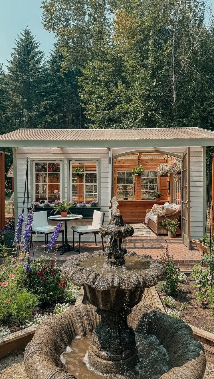 Dream Garden for Your Aesthetic: 20 Ideas to Steal 18 A stone tiered fountain sits before a cozy white garden shed with an outdoor seating area.