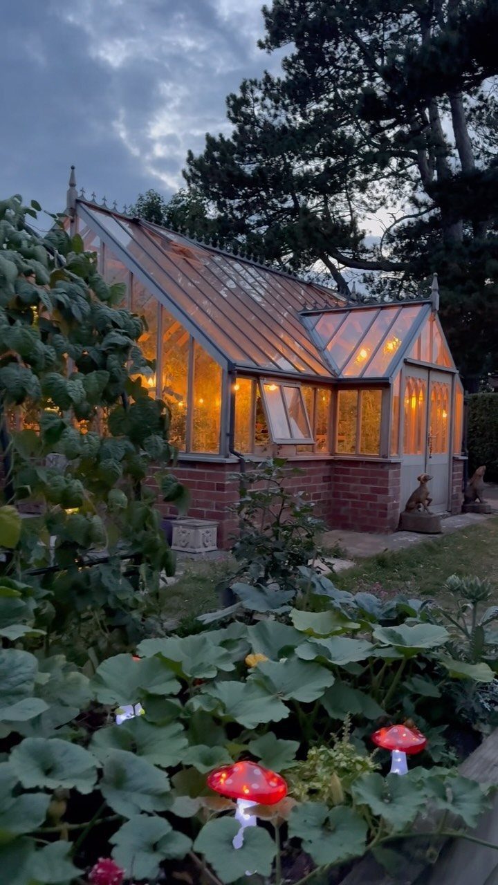Dream Garden for Your Aesthetic: 20 Ideas to Steal 8 A glowing glass greenhouse with a brick foundation surrounded by lush green garden plants at dusk.
