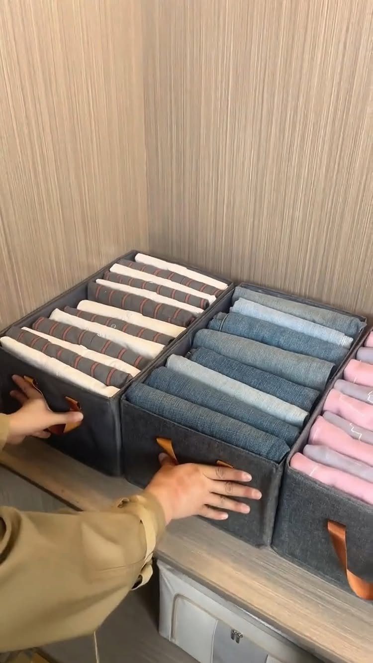 Grey fabric bins containing clothes folded vertically on a wooden shelf.