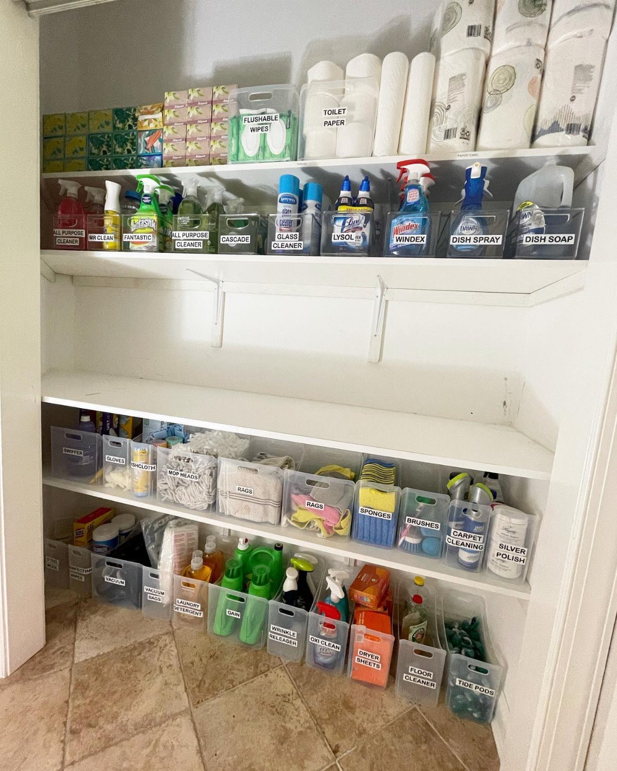 Cleaning supplies organized in clear plastic bins on white closet shelves with labels.