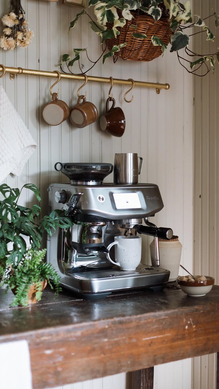 16 Farmhouse Coffee Bar Ideas for Pantry Nooks to Pallet Walls 16 An espresso machine on a wooden counter with hanging mugs and greenery in a farmhouse setting.