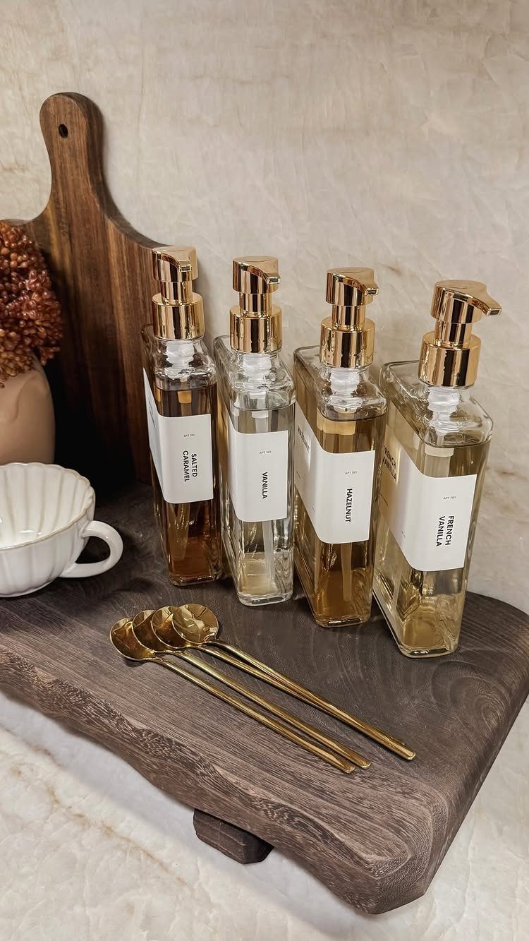 16 Farmhouse Coffee Bar Ideas for Pantry Nooks to Pallet Walls 2 Four glass pump bottles with gold tops and three gold spoons on a wooden tray.
