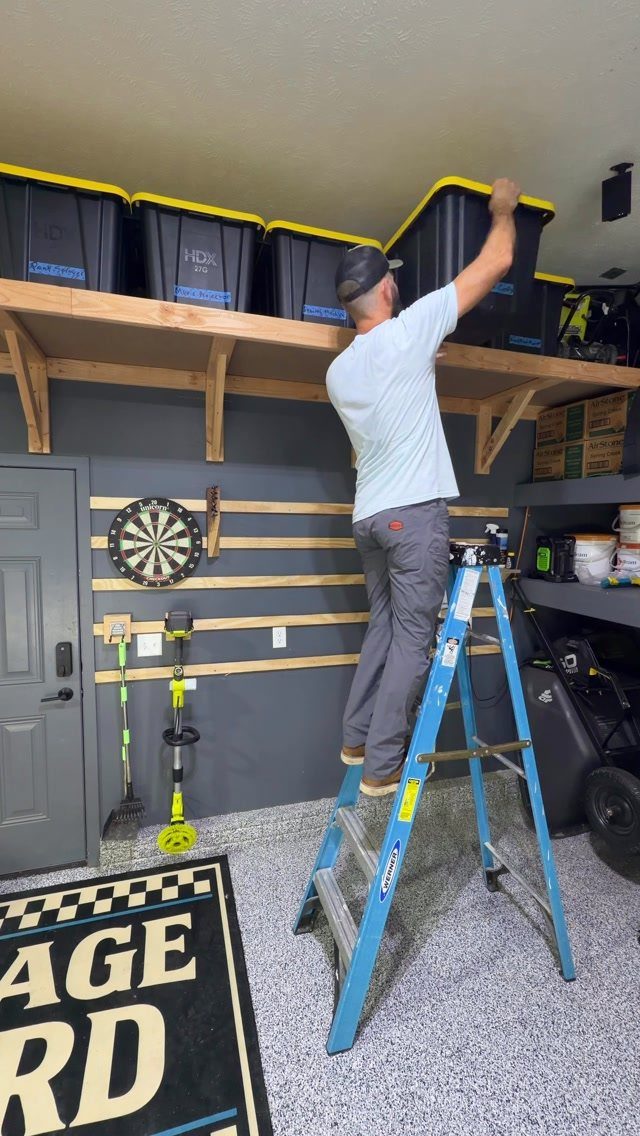 Man on a blue ladder reaching for black storage bins on a high wooden garage shelf.