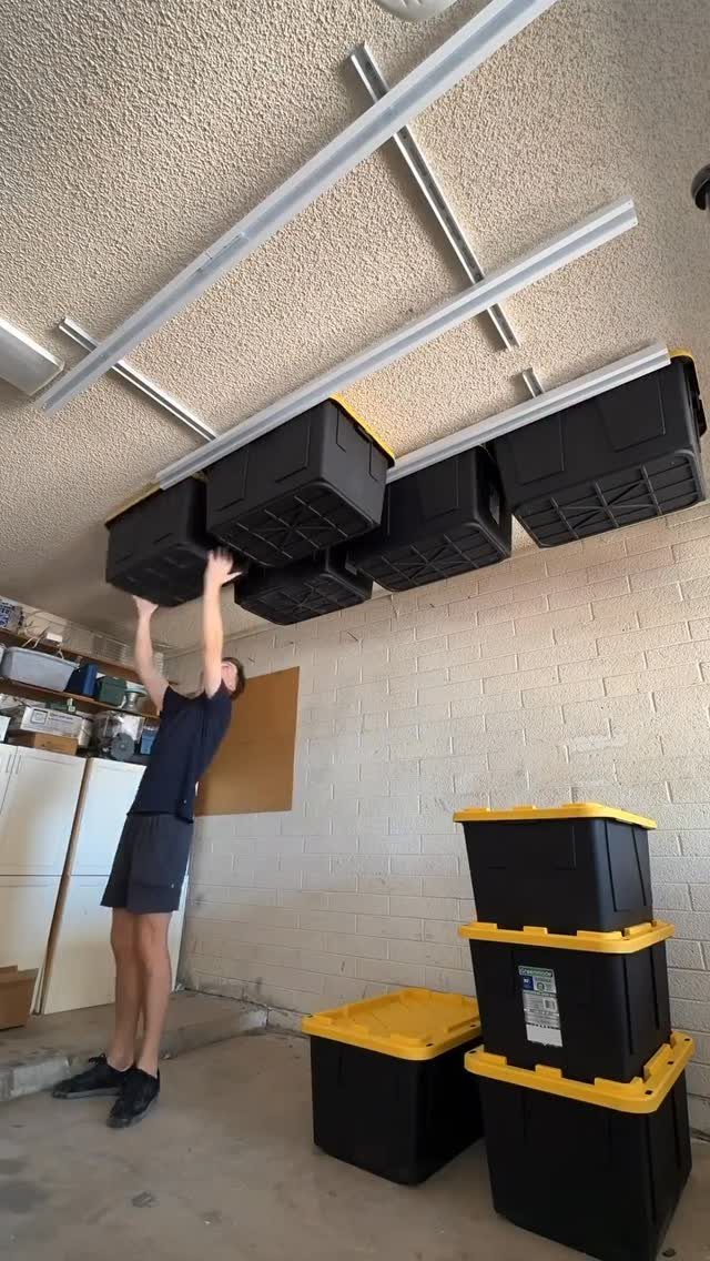 A person lifting a black storage bin from a white overhead rack in a garage.
