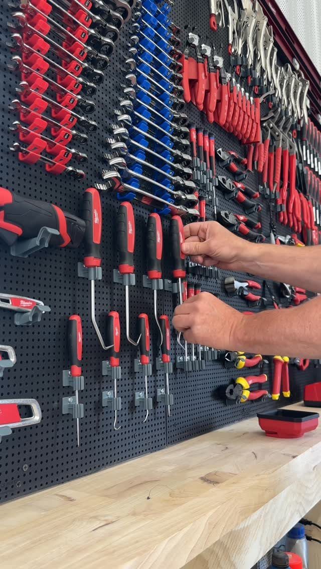 Hands reaching for red and black hand tools organized on a black pegboard above a wooden workbench.