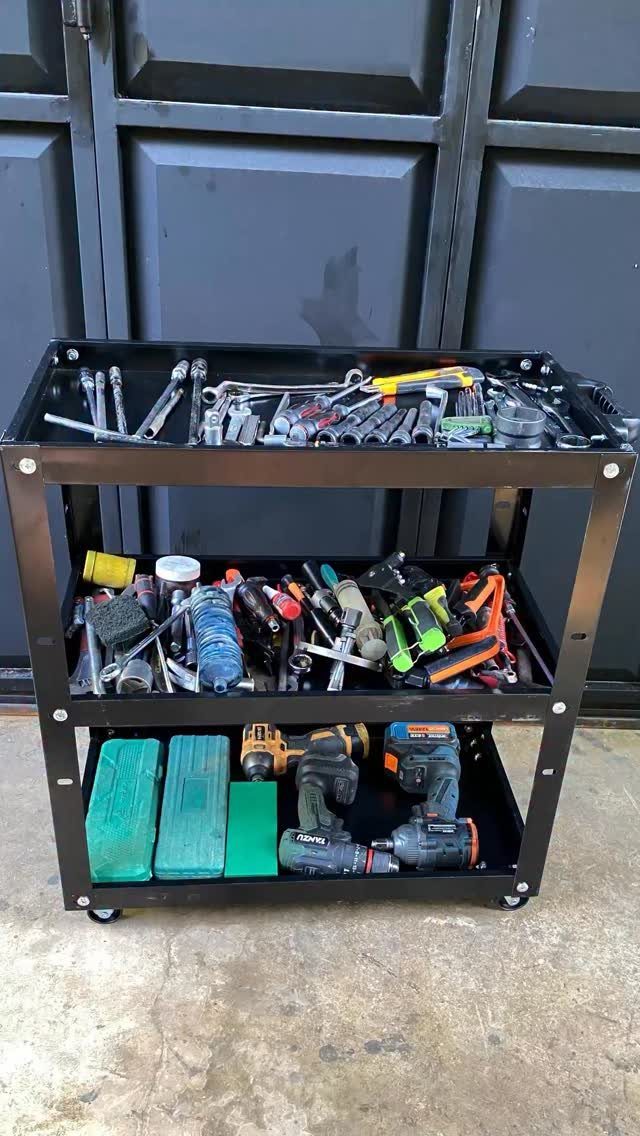 A black three-tier rolling cart filled with various hand tools and power drills on concrete.