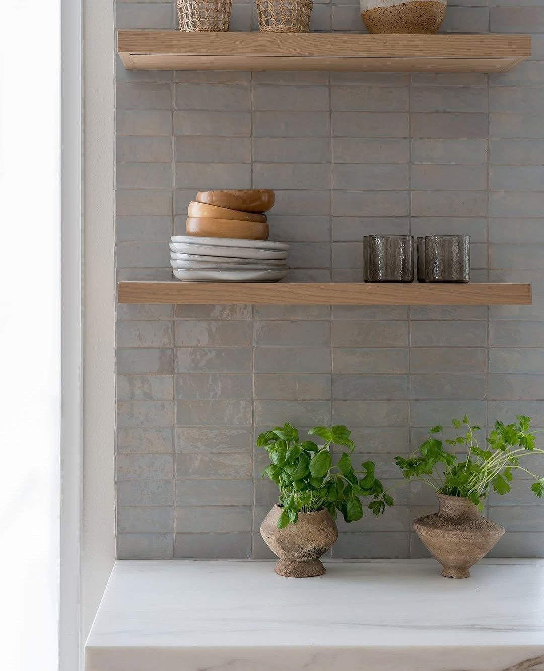 Grey blue tiled kitchen backsplash with wooden floating shelves and small potted plants on marble countertop.
