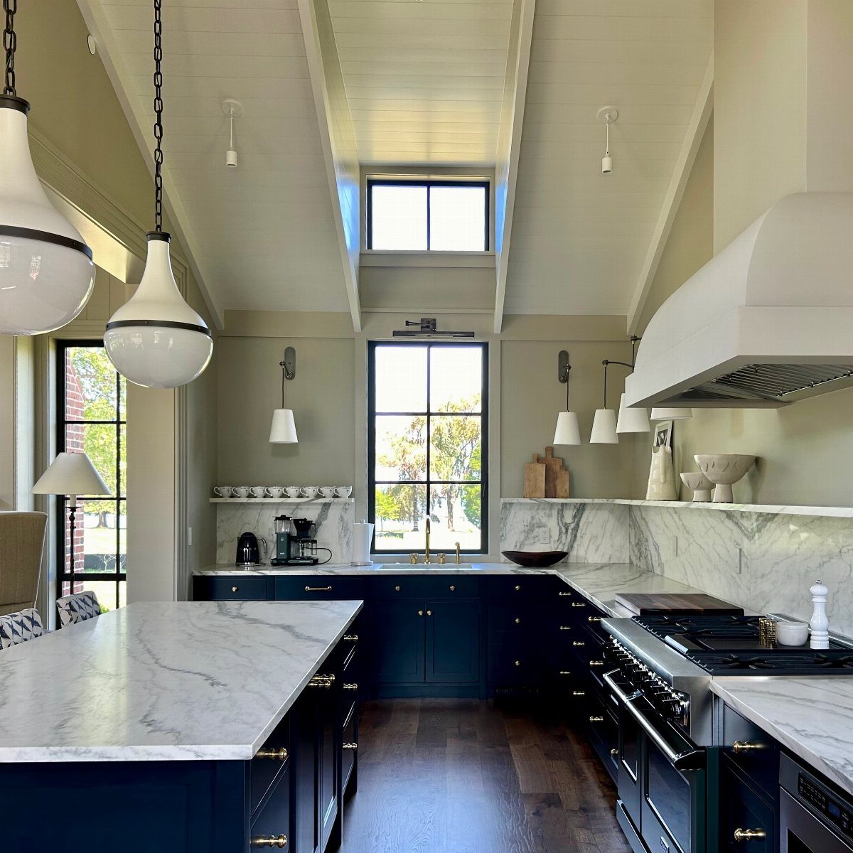 Navy blue kitchen cabinets with white marble countertops and large black-framed windows.