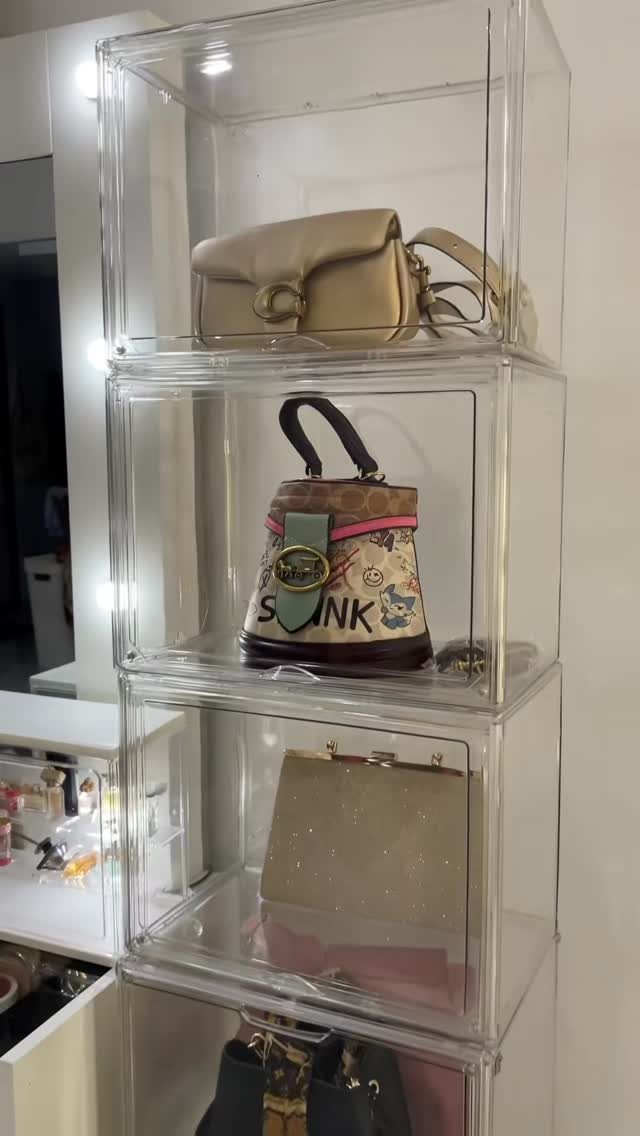 Clear acrylic storage bins stacked vertically containing various designer handbags on a white wall.
