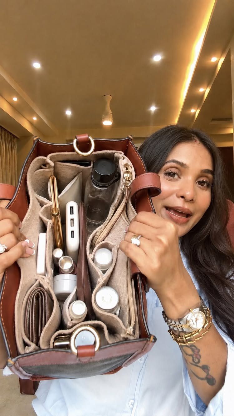 A woman holds open a brown leather handbag showing organized compartments with various personal items inside.