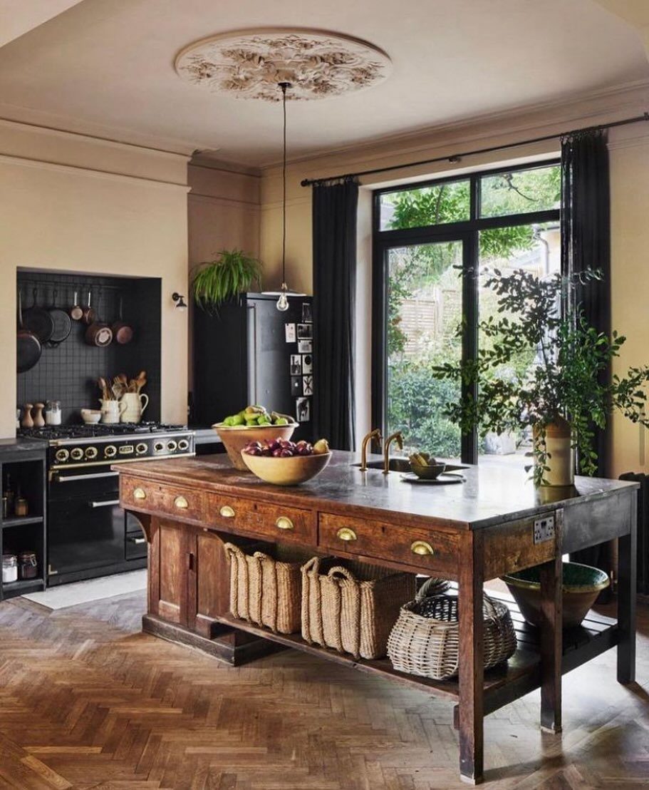 A large reclaimed wood kitchen island with wicker baskets sits on herringbone wood flooring.