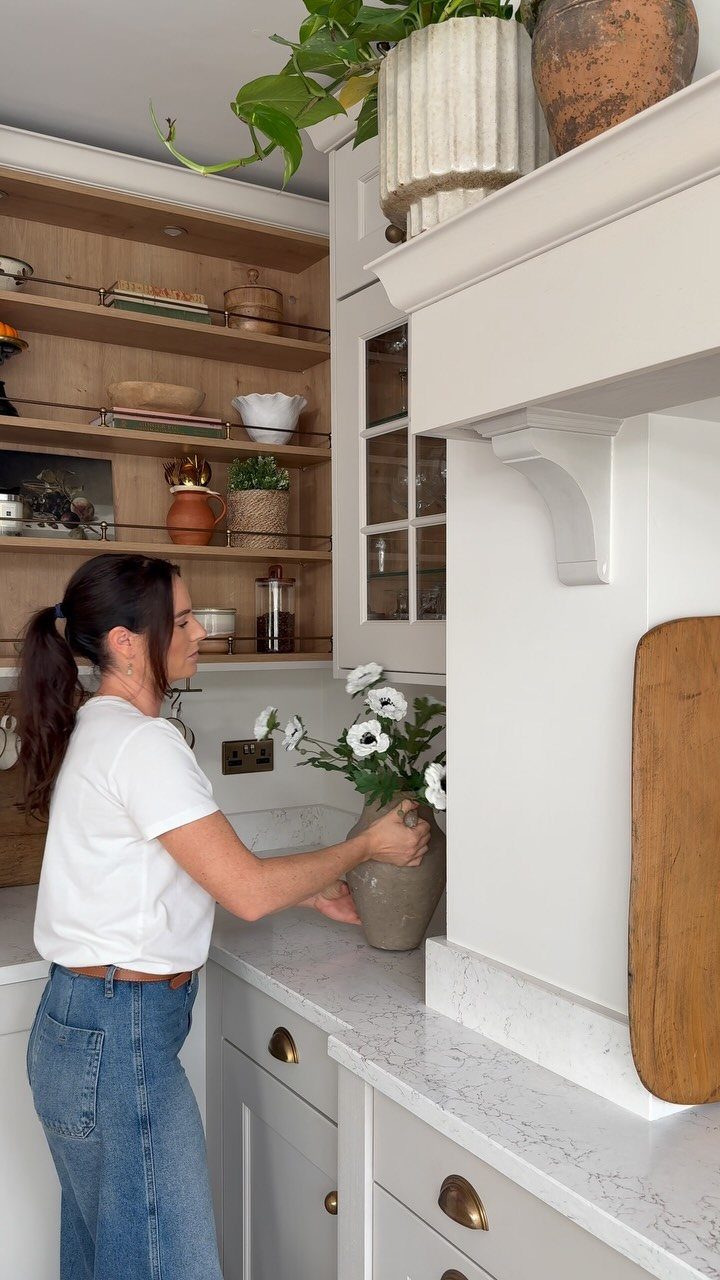 Woman placing a vase of white flowers on a marble kitchen countertop near open shelving.