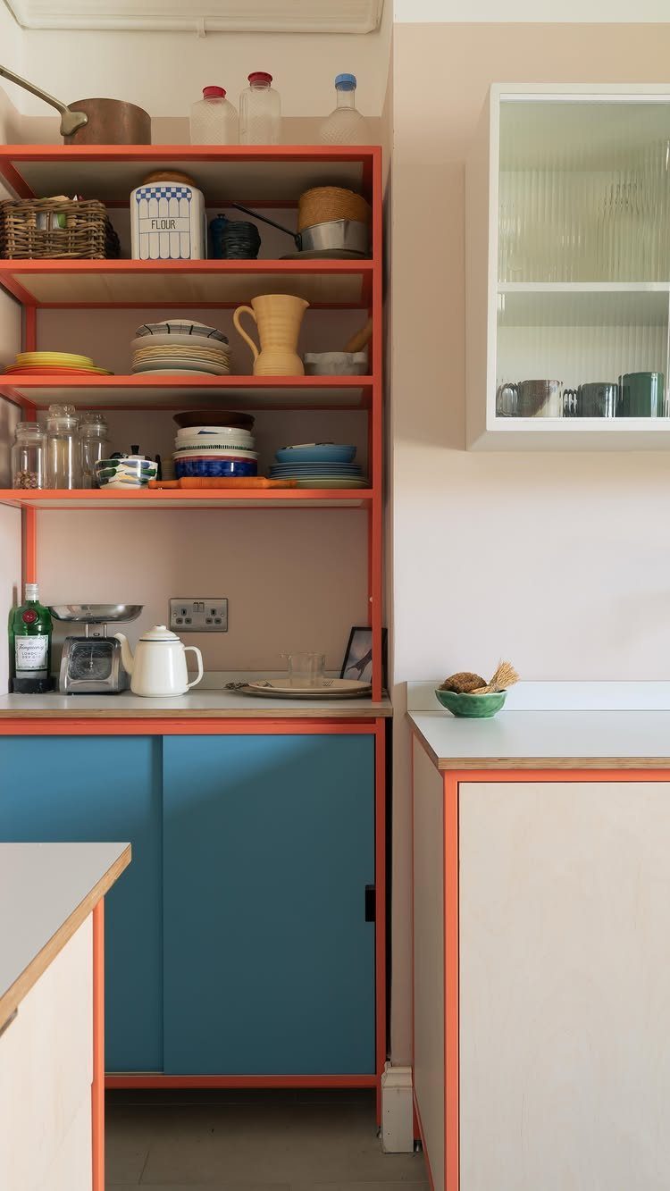 Orange kitchen shelving and blue cabinets holding various dishes and kitchenware against a light wall.