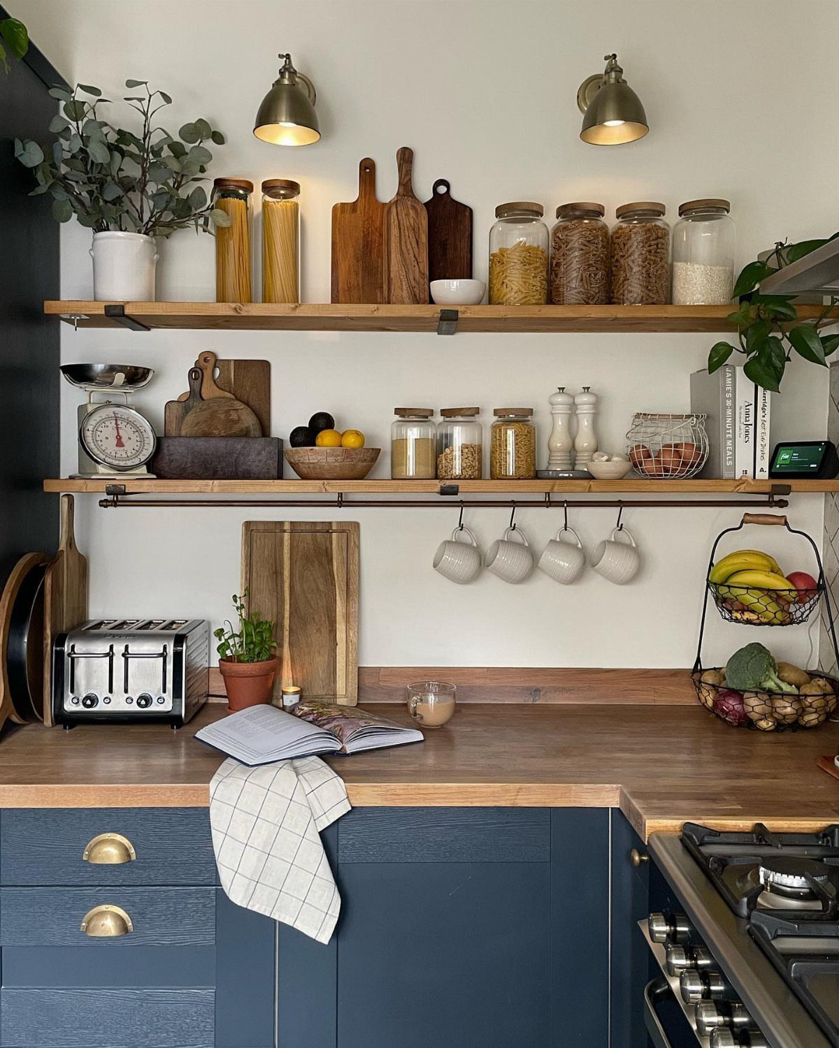 Wooden floating shelves holding glass jars, cutting boards, and hanging mugs in a modern kitchen.