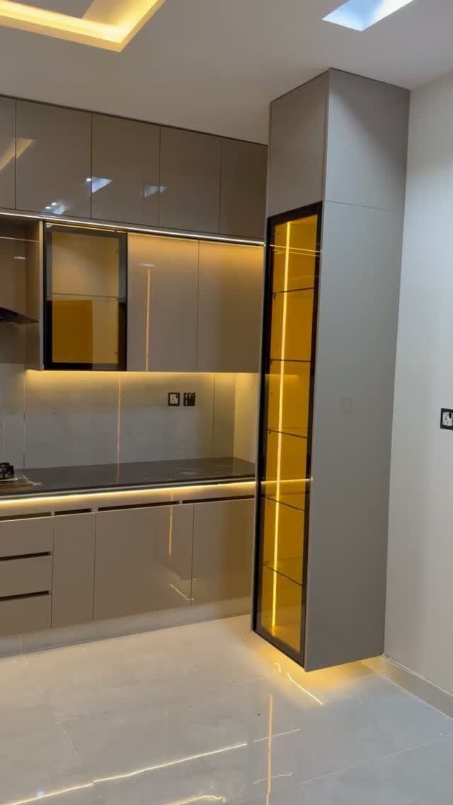 Modern straight kitchen with glossy taupe cabinets, dark countertops, and illuminated glass pantry shelves.