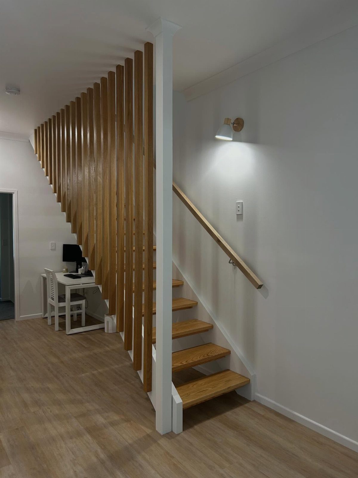 A small white desk and computer monitor located under a wooden slatted staircase.