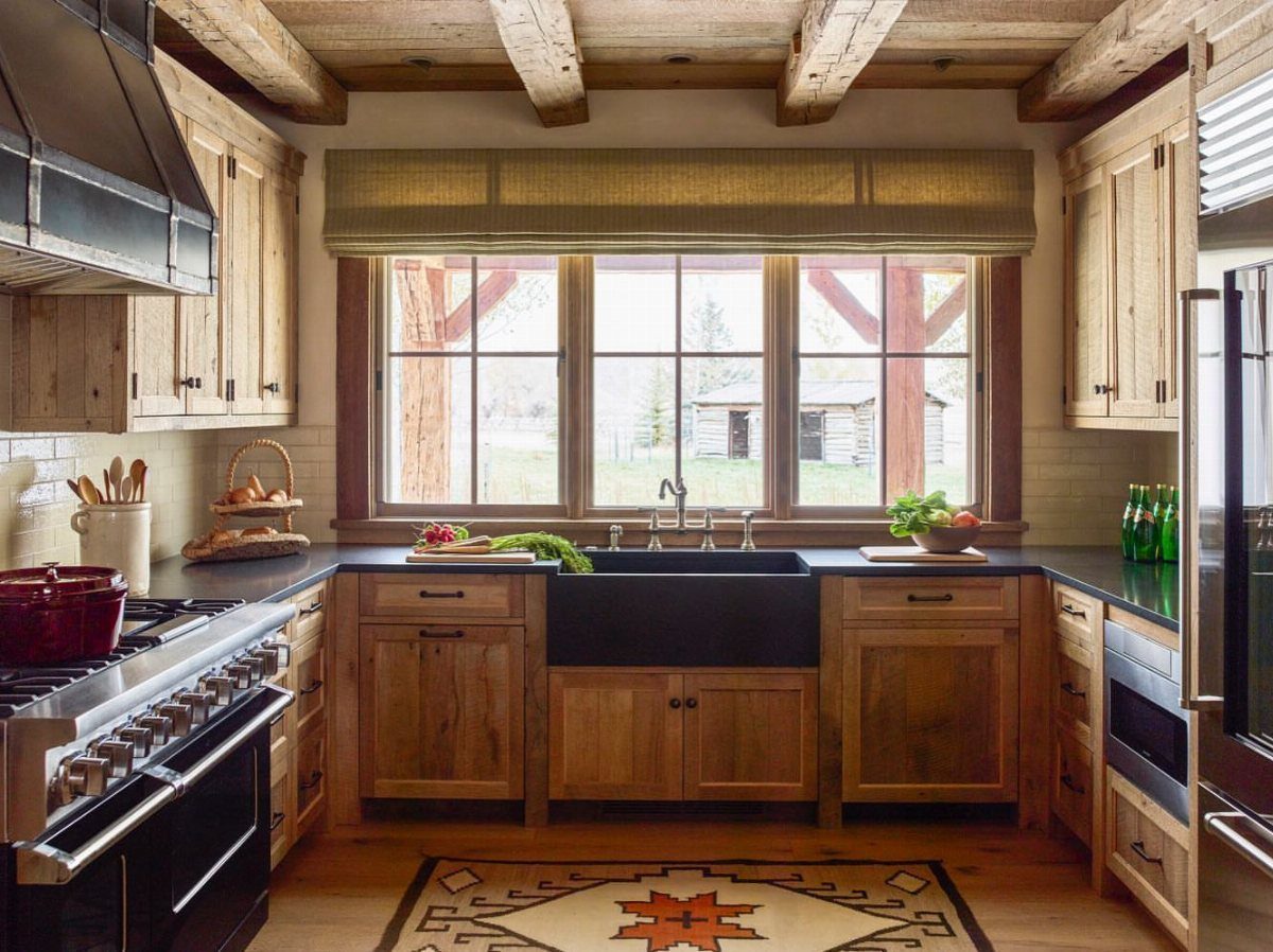 Reclaimed wood kitchen cabinets with dark countertops and a black farmhouse sink under large windows.