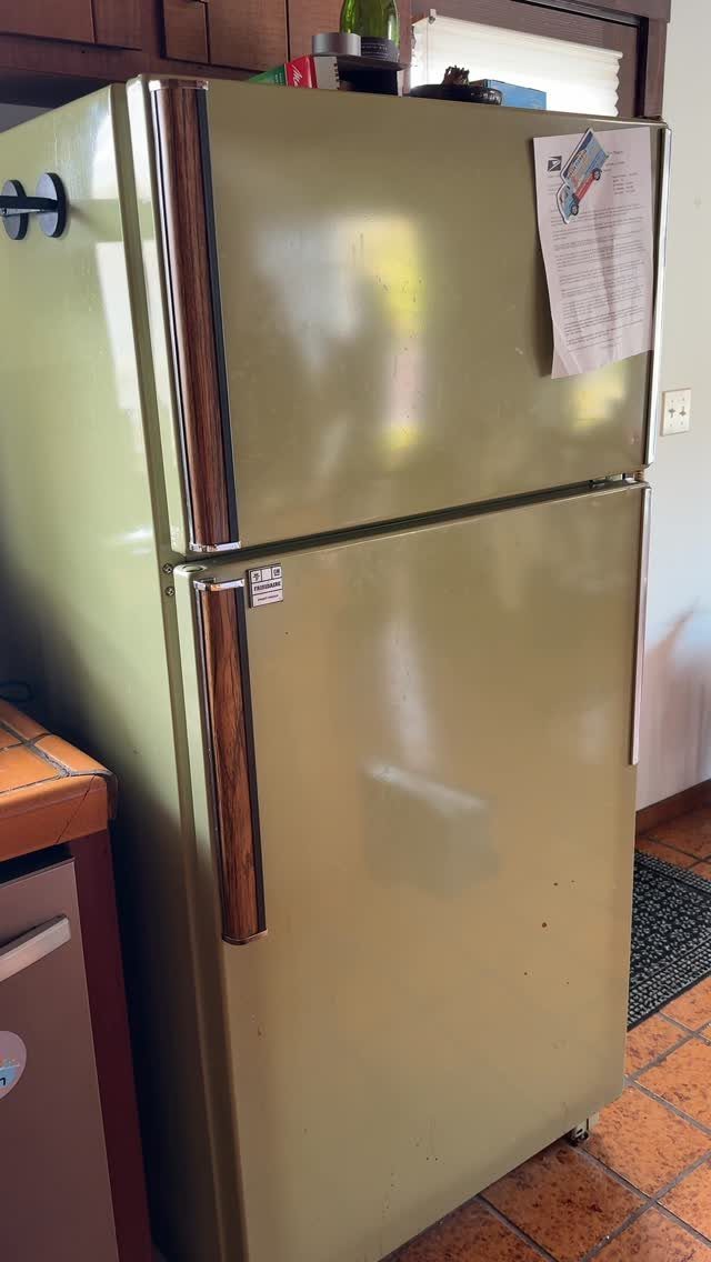 An avocado green retro-style refrigerator stands on a terracotta tile kitchen floor.