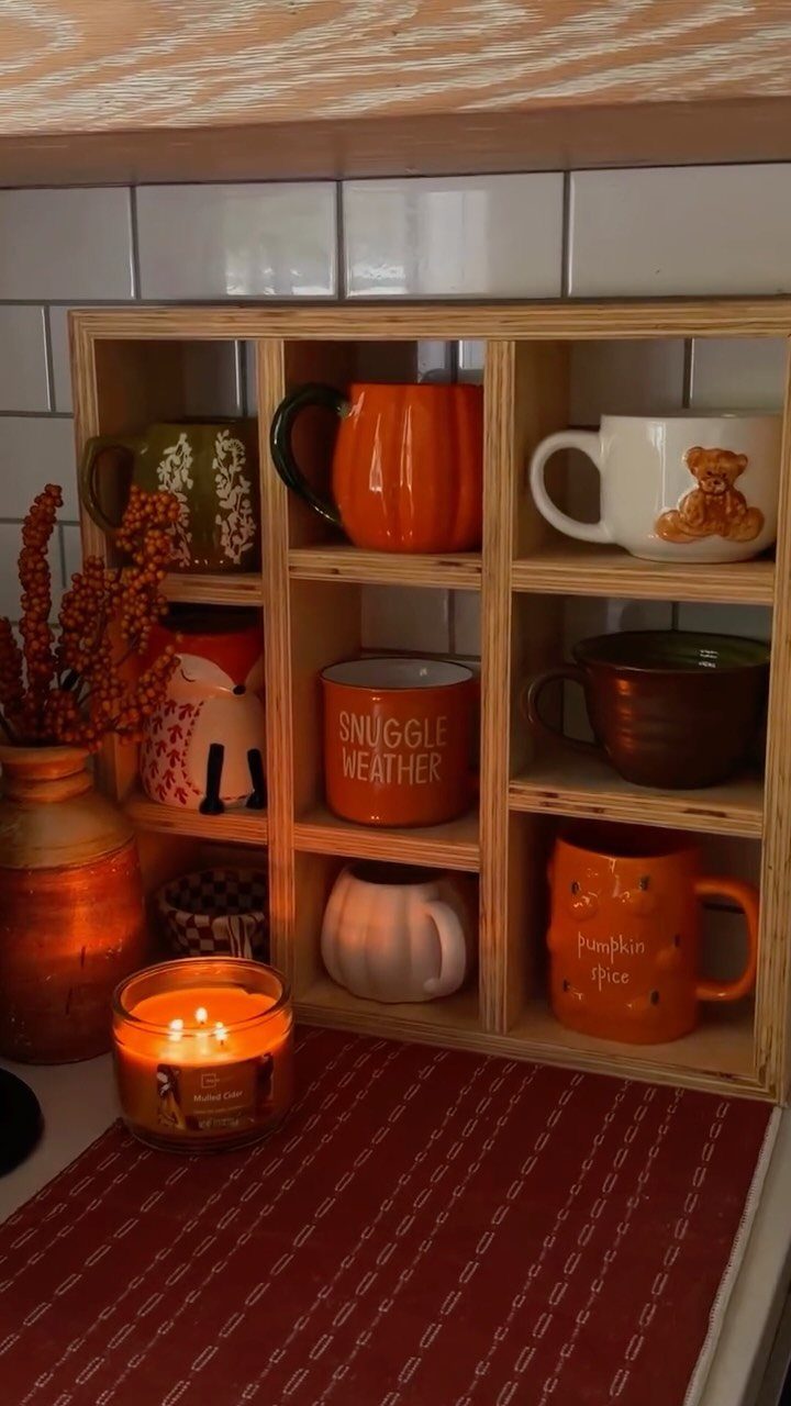 Seasonal coffee mugs in a wooden cubby shelf next to a lit decorative candle.