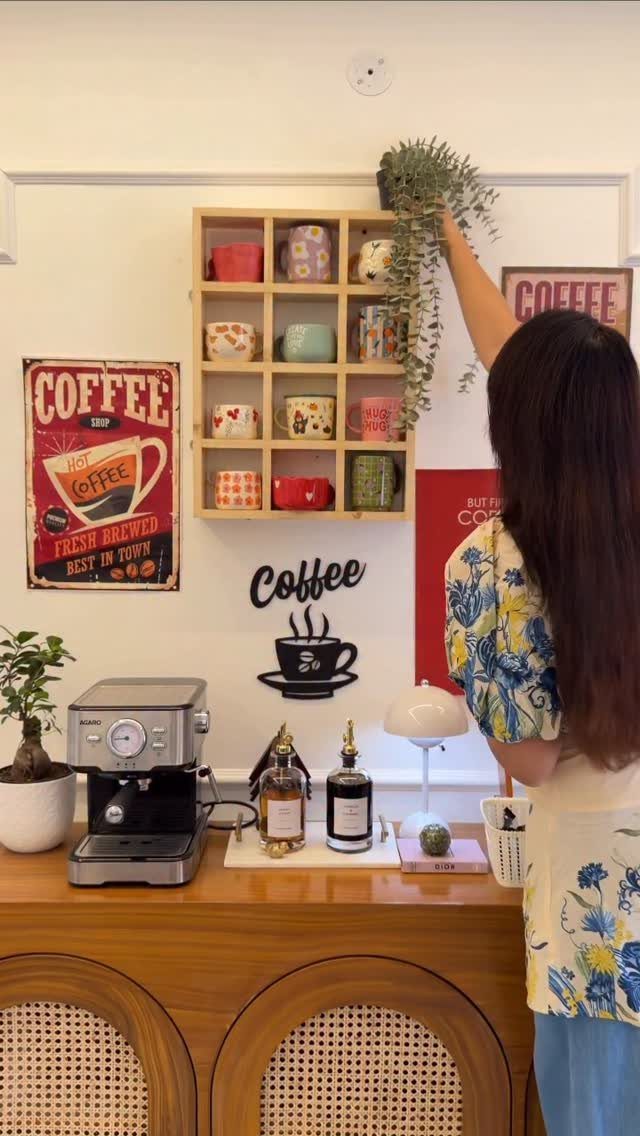 A woman arranging a plant on a wooden cubby shelf filled with colorful mugs above a coffee station.